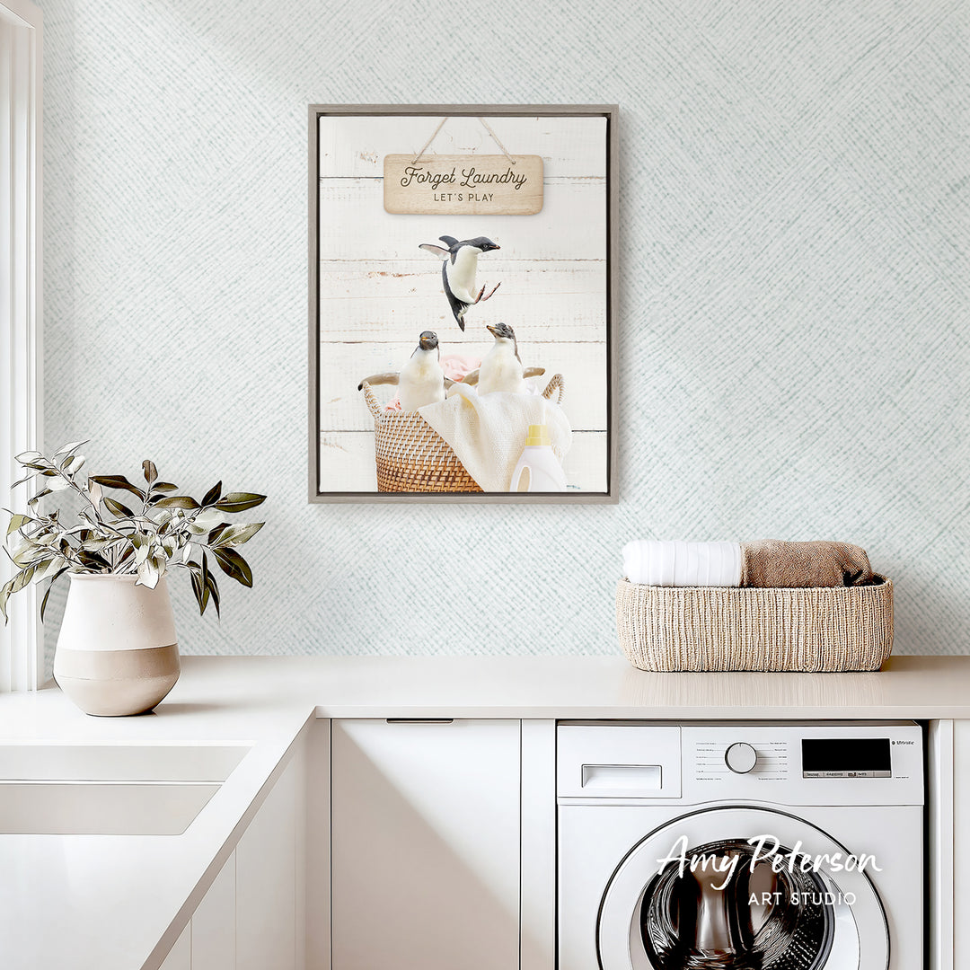 a framed print of penguins in a basket, hanging on a wall above a white countertop with a plant and a basket of towels.