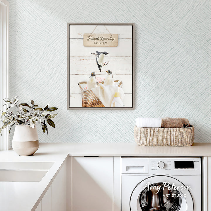 a framed print of penguins in a basket, hanging on a wall above a white countertop with a plant and a basket of towels.