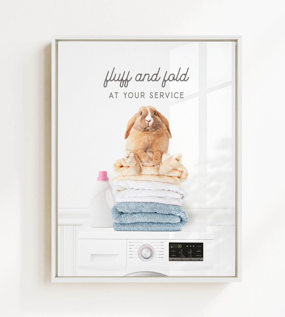 A framed image depicting a fluffy brown rabbit sitting atop a stack of folded towels, with a white washing machine and detergent bottle in the background.