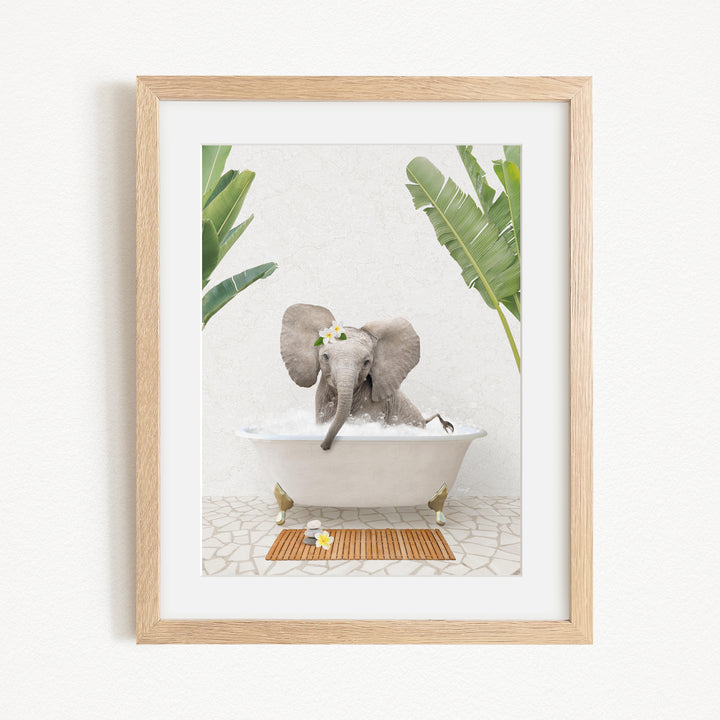 A gray elephant is sitting in a white bathtub, surrounded by green leaves, with a wooden mat on the floor in front of the tub.