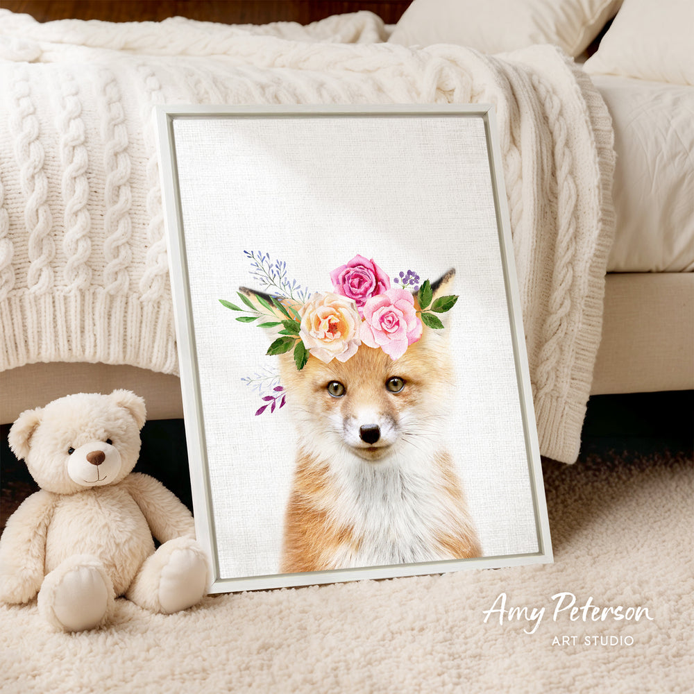 A framed print of a fox wearing a flower crown is displayed on a white bedspread, accompanied by a teddy bear on the floor.