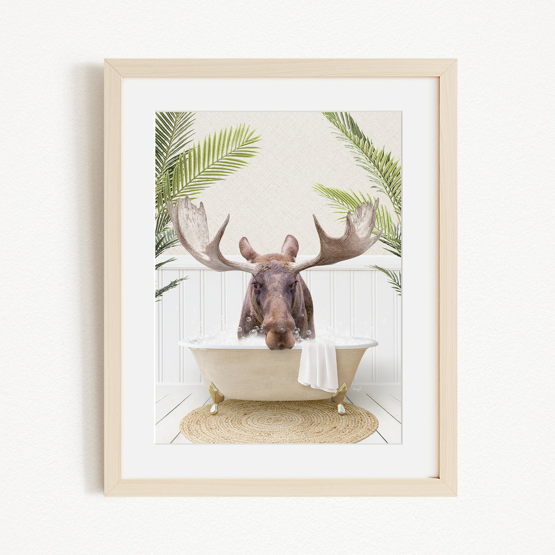 A framed image depicting a moose with large antlers sitting in a bathtub, surrounded by tropical plants.