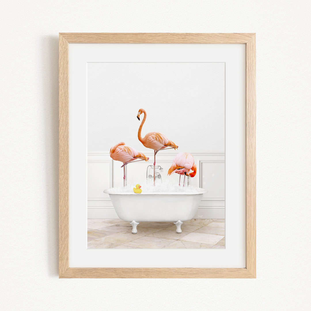 A framed image depicting three flamingos standing in a white bathtub filled with water, with a yellow rubber duck floating in the water.