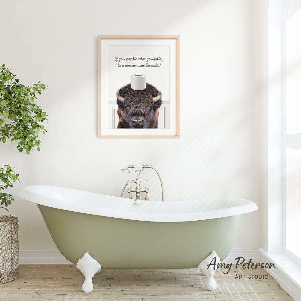 a bathroom with a green clawfoot bathtub, a framed picture of a bison with a toilet paper roll on its head, and a potted plant on the floor.