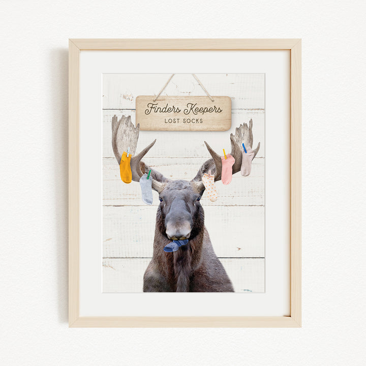 A framed image of a moose with a sign that says "Finders Keepers" hanging above it, along with several pairs of socks hanging from its antlers.