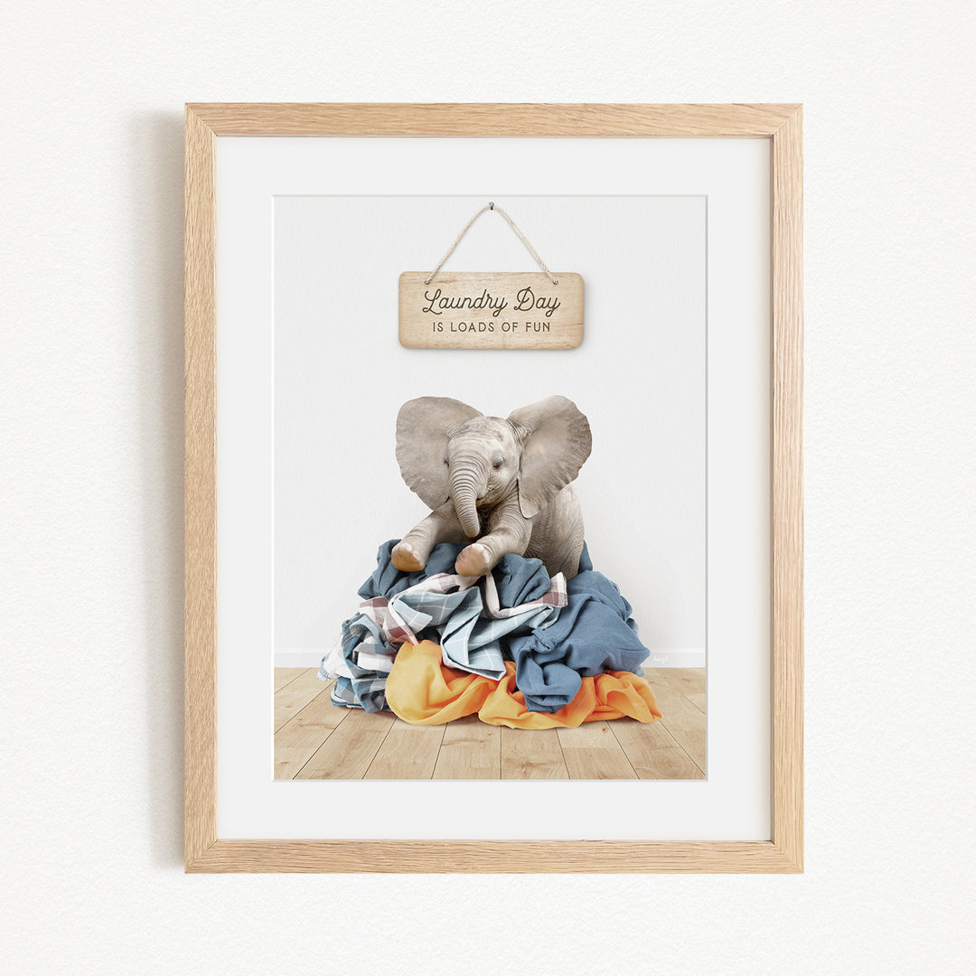 A framed image depicting a baby elephant sitting atop a pile of laundry, with a sign above that reads "Laundry Day is loads of fun".