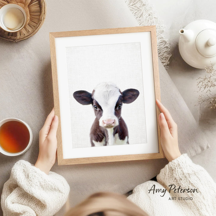 A person is holding a framed picture of a black and white cow, with a teapot, a cup of tea, and a bowl of tea visible in the background.