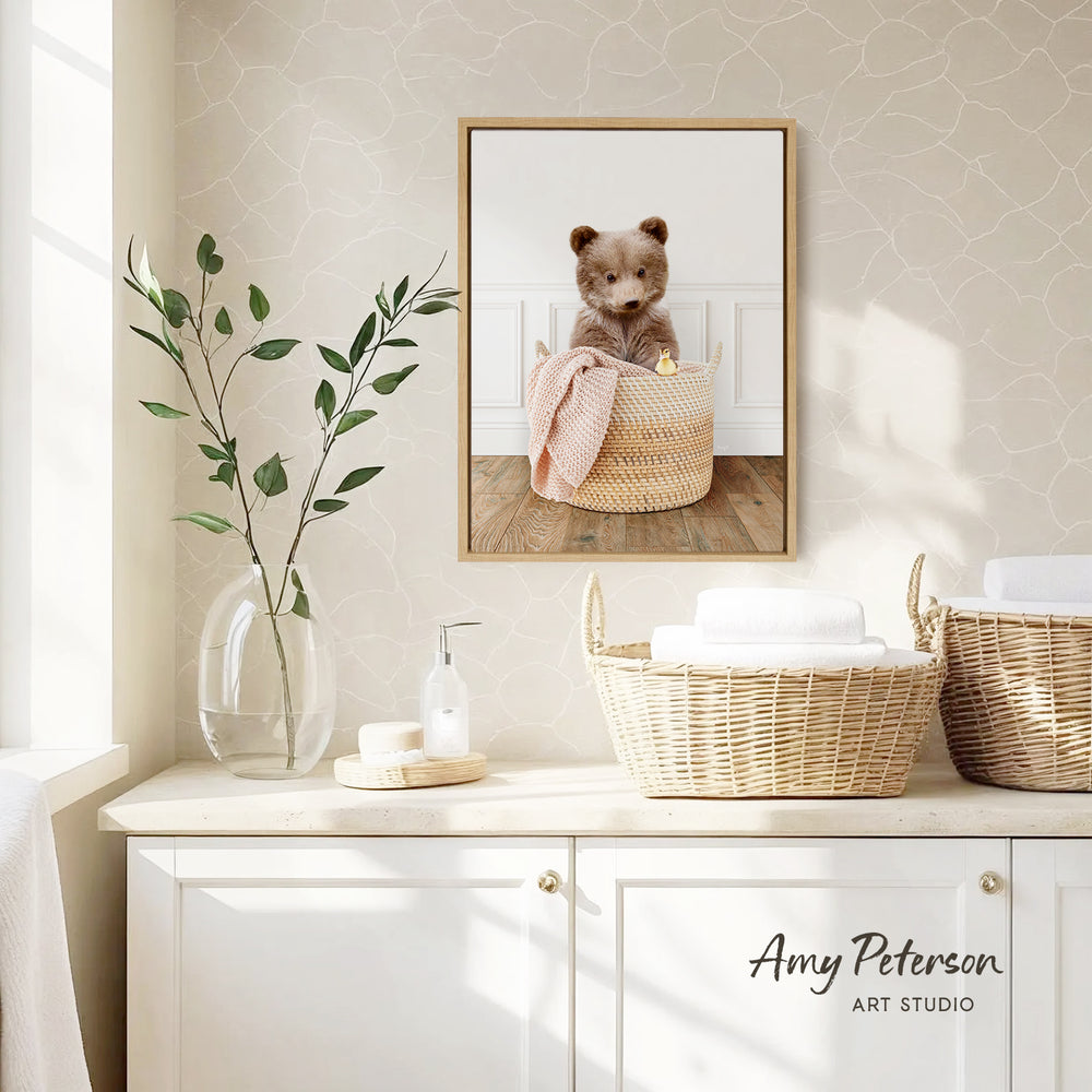 a cozy bathroom scene with a framed photo of a bear sitting in a basket, a vase with green leaves, and baskets on a counter.