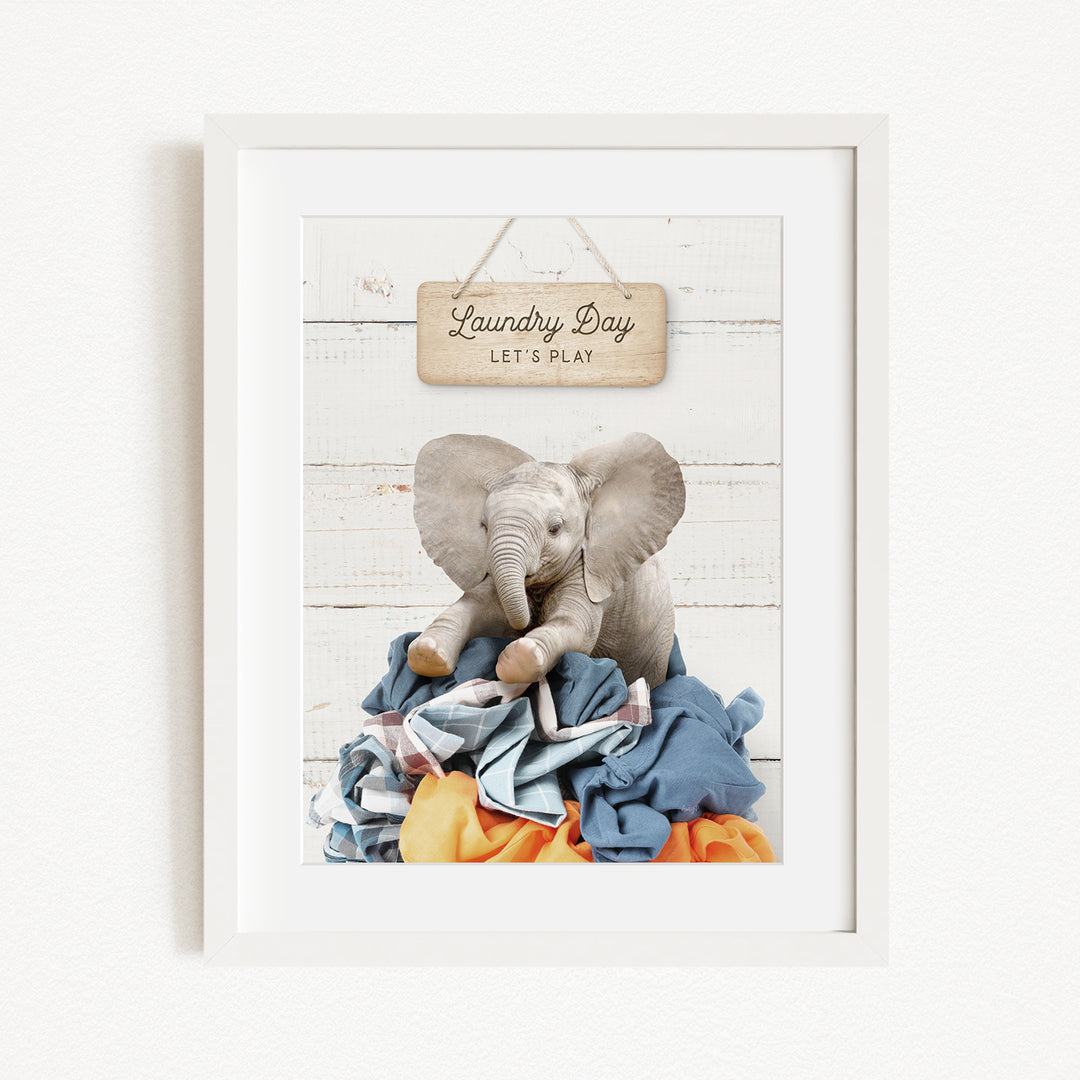 A framed image of a stuffed elephant sitting atop a pile of laundry, with a wooden sign above that says "Laundry Day Let's Play".