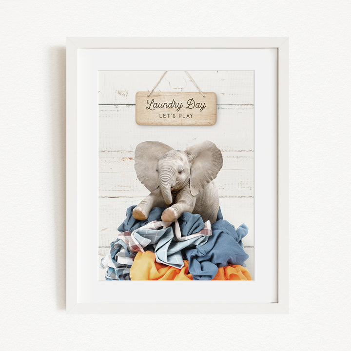 A framed image of a stuffed elephant sitting atop a pile of laundry, with a wooden sign above that says "Laundry Day Let's Play".