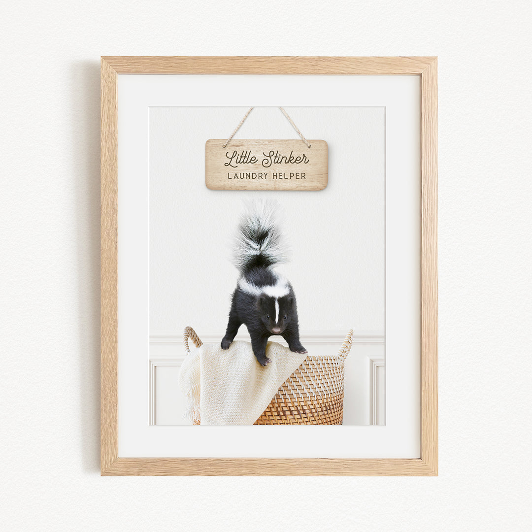 A framed photograph of a black and white skunk standing on a woven basket, with a wooden sign above it that reads "Little Skinker Laundry Helper".