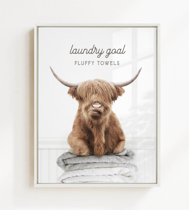 A brown cow with long horns is sitting on a stack of fluffy towels, with the text "laundry goal" and "FLUFFY TOWELS" displayed above the image.