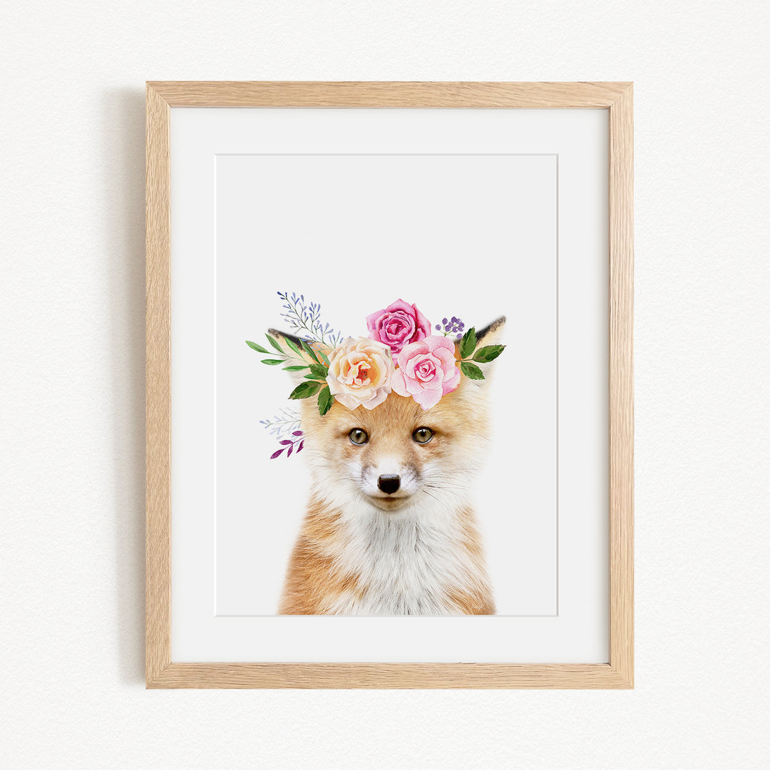 A framed print of a fox wearing a floral crown of pink and white roses and greenery.