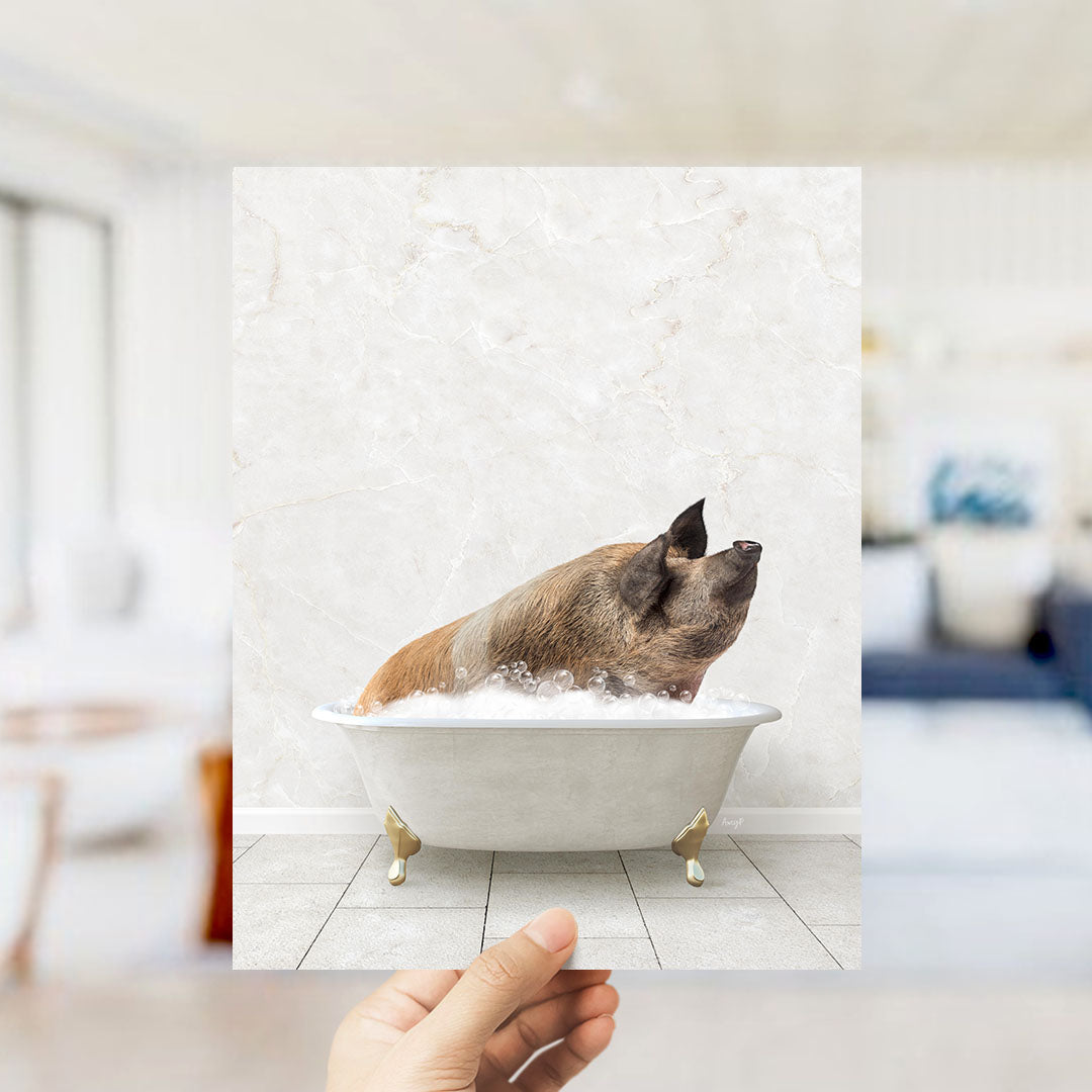 A hand holds up a framed image of a pig sitting in a bathtub filled with bubbles.