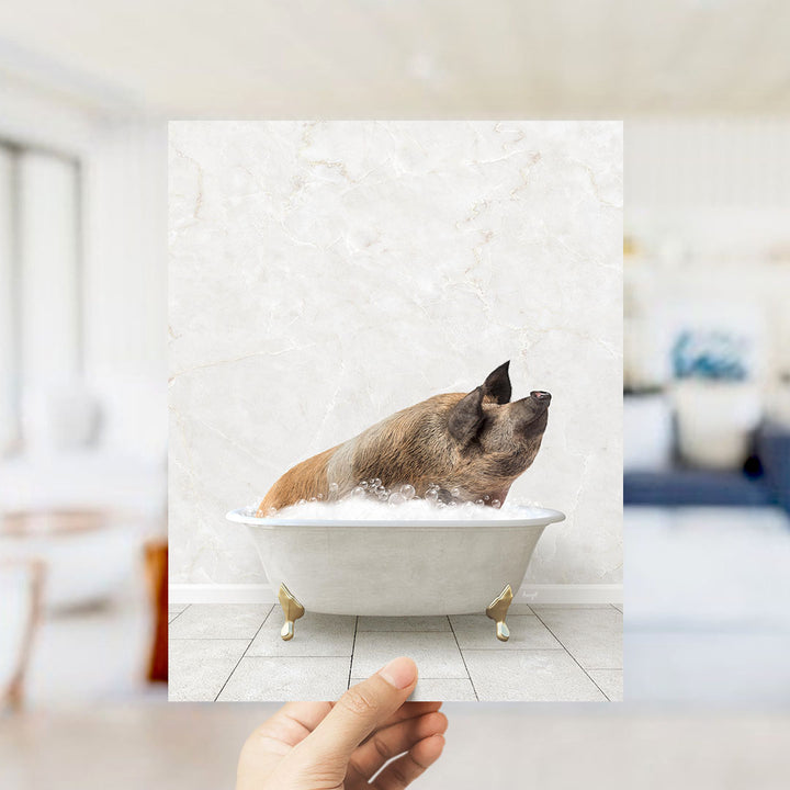 A hand holds up a framed image of a pig sitting in a bathtub filled with bubbles.