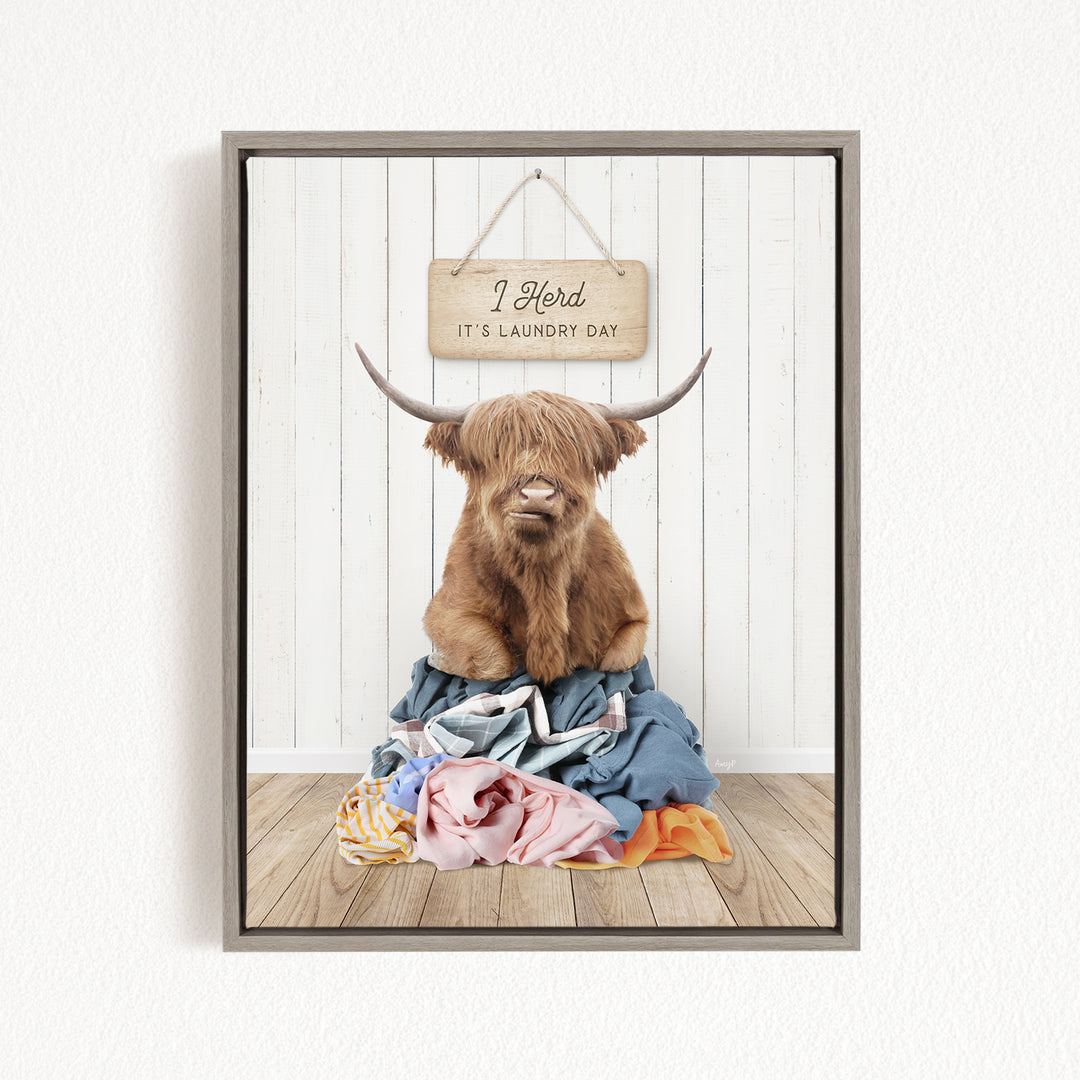 A framed image of a Highland cow with large horns sitting on a pile of laundry, with a sign above it that says "I herd it's laundry day".