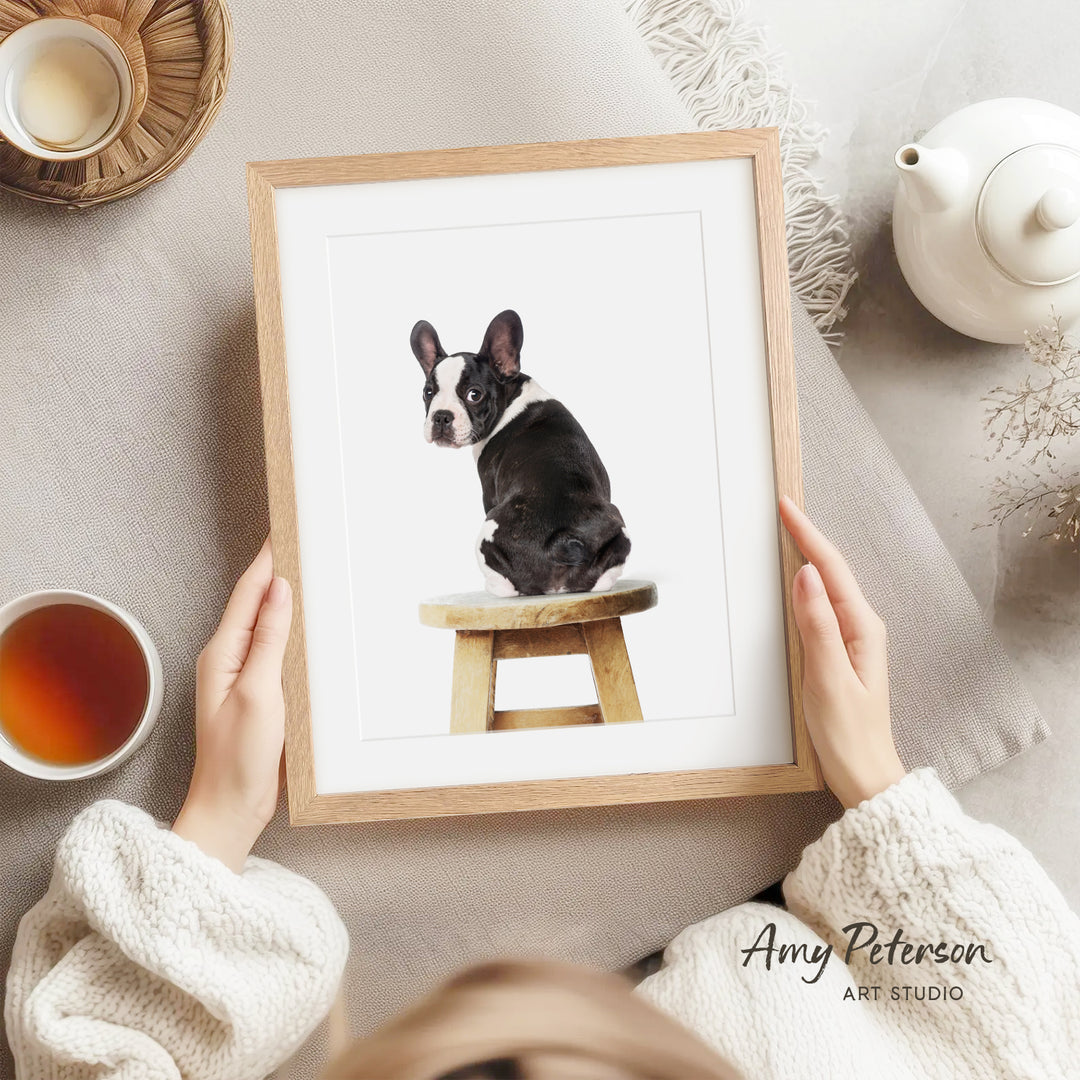 A person is holding a framed photograph of a black and white dog sitting on a wooden stool, with a teapot and a cup of tea nearby.