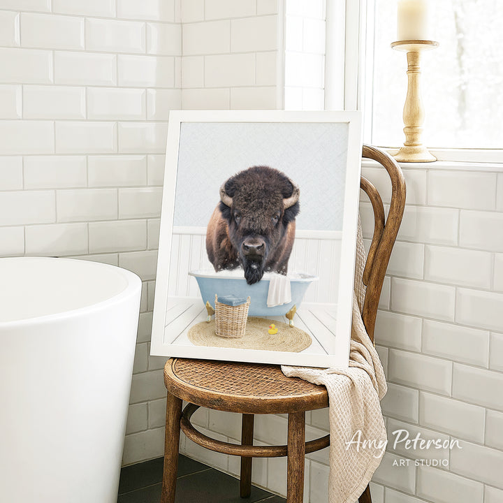 A framed image of a buffalo taking a bath in a tub, with a towel and basket nearby, is displayed on a wooden chair in a bathroom setting.
