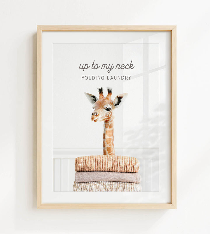 A framed image of a giraffe standing on a pile of clothes, with the text "up to my neck" and "Folding Laundry" written above it.