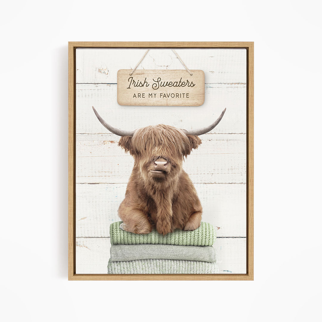 A brown Highland cow with large horns is sitting on a stack of green and gray blankets, with a sign above that says "Irish Sweaters are my favorite".
