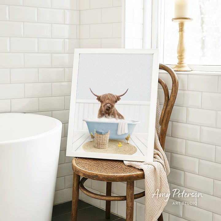 A framed image of a Highland cow sitting in a bathtub, with a towel and a basket nearby, is displayed on a wooden chair in a bathroom setting.