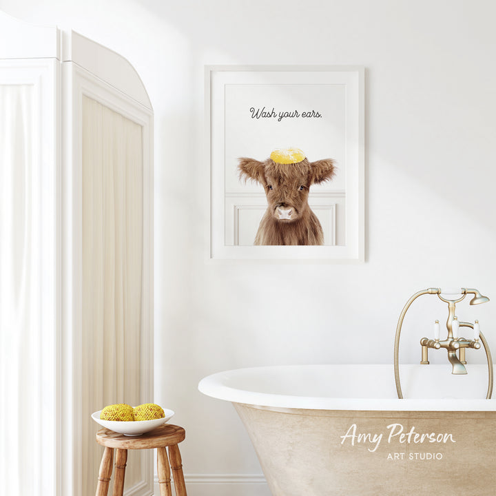 a bathroom with a framed picture of a cow on the wall, a wooden stool, a bowl of fruit, and a bathtub with a faucet.