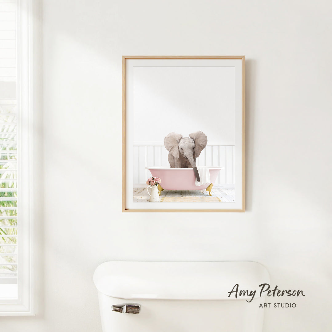 A framed image of an elephant sitting in a pink bathtub is hanging on a white wall in a bathroom.