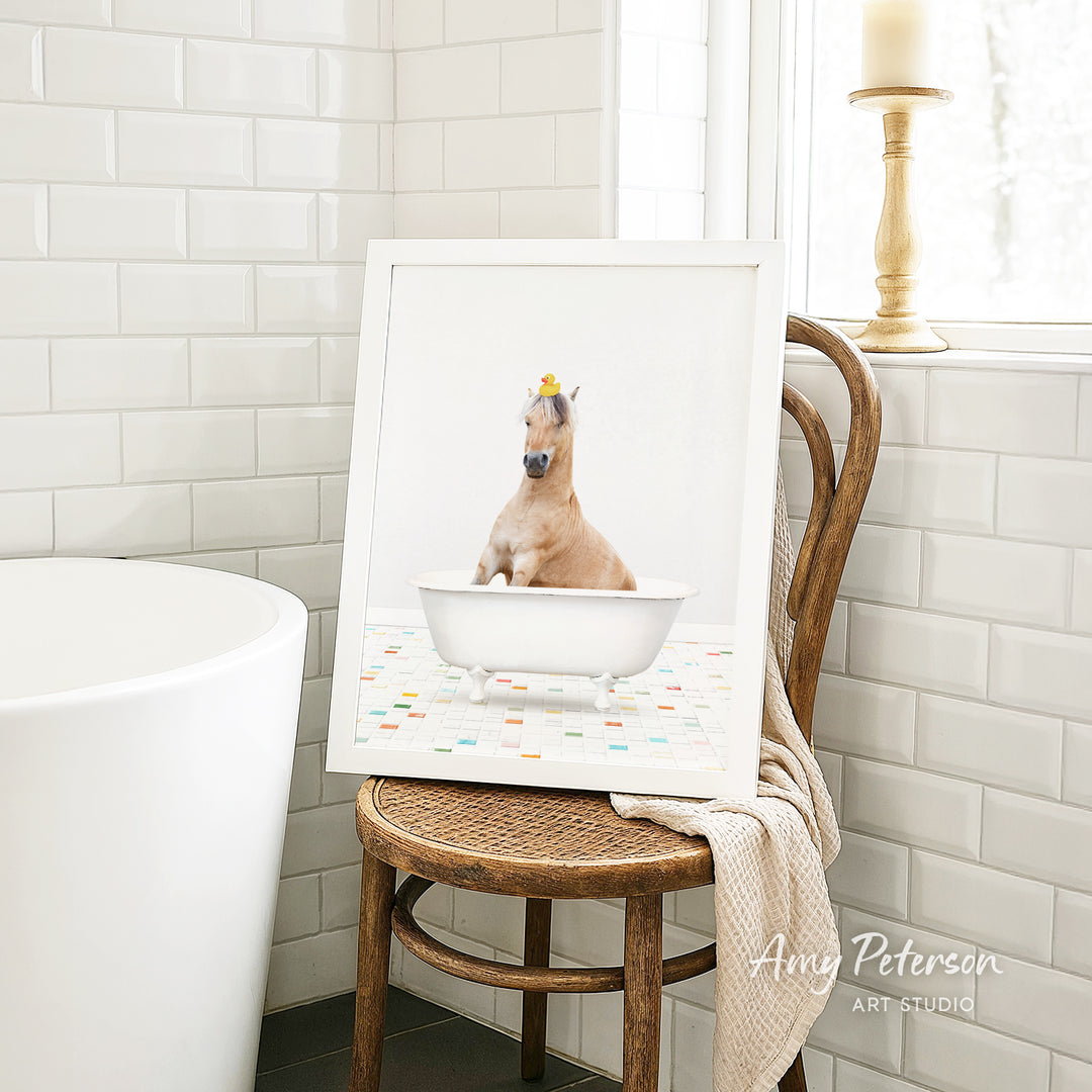 A framed image of a horse sitting in a bathtub, with a towel draped over the back of a wooden chair in a bathroom setting.