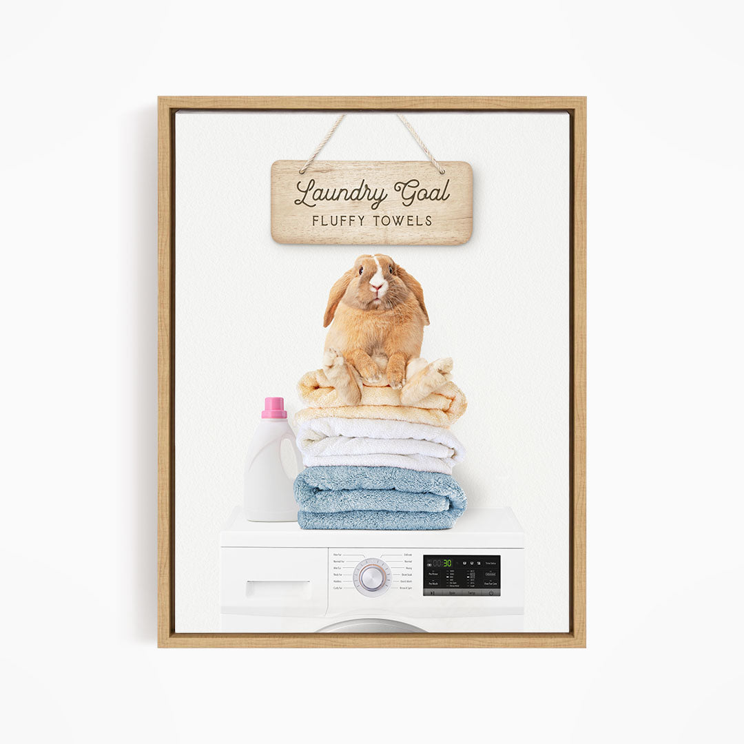 A wooden sign with the text "Laundry Goal Fluffy Towels" hangs above a white washing machine with a pile of folded towels on top.