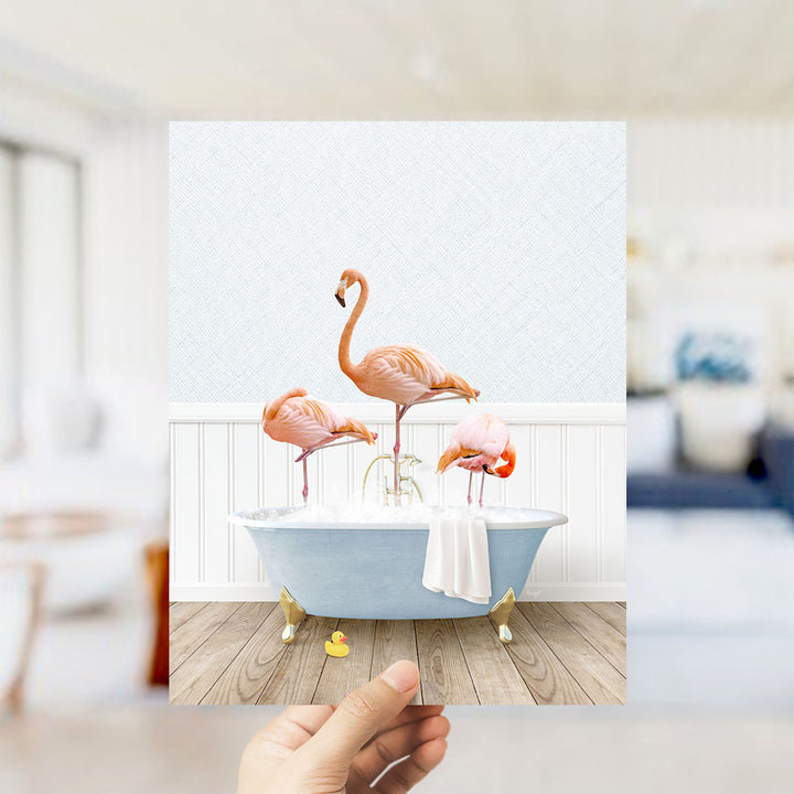 A hand holds up a framed image depicting three flamingos standing in a bathtub filled with bubbles.