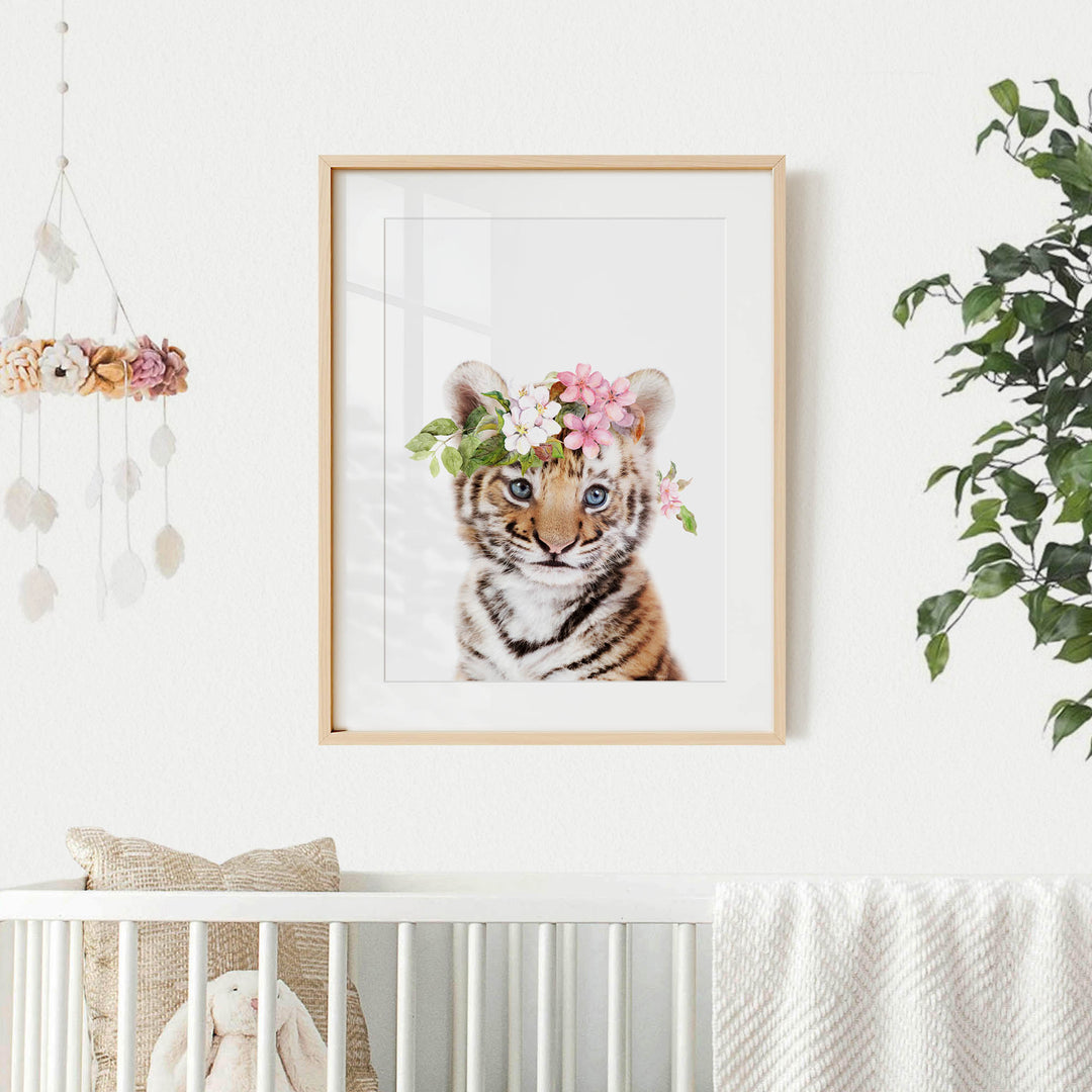 A framed photograph of a tiger cub wearing a floral crown is displayed on a white wall, with a hanging mobile and a plant in the background.