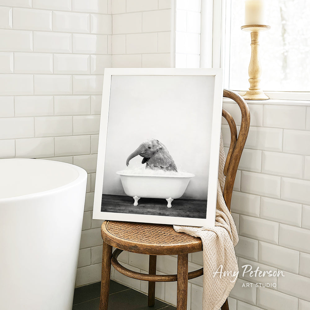 A framed black and white photograph of an elephant sitting in a bathtub, with a towel draped over the back of a wooden chair in a bathroom setting.