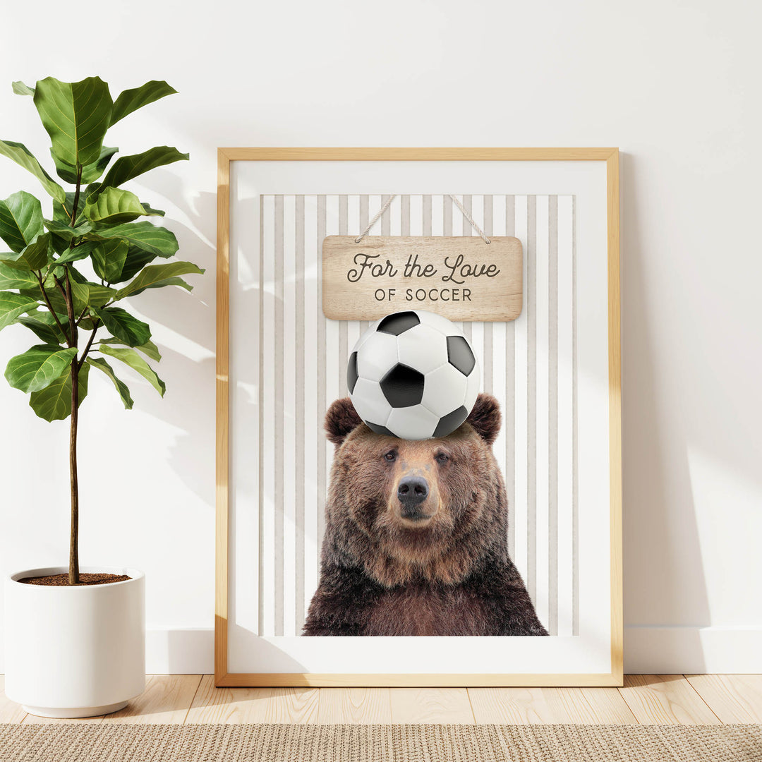 A framed image of a brown bear with a soccer ball on its head, accompanied by a wooden sign that says "For the Love of Soccer".