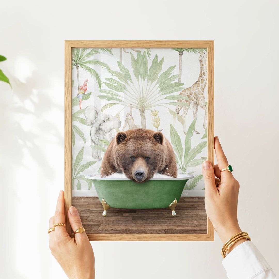 A person's hands are holding a framed picture of a brown bear sitting in a green bathtub, surrounded by tropical foliage.