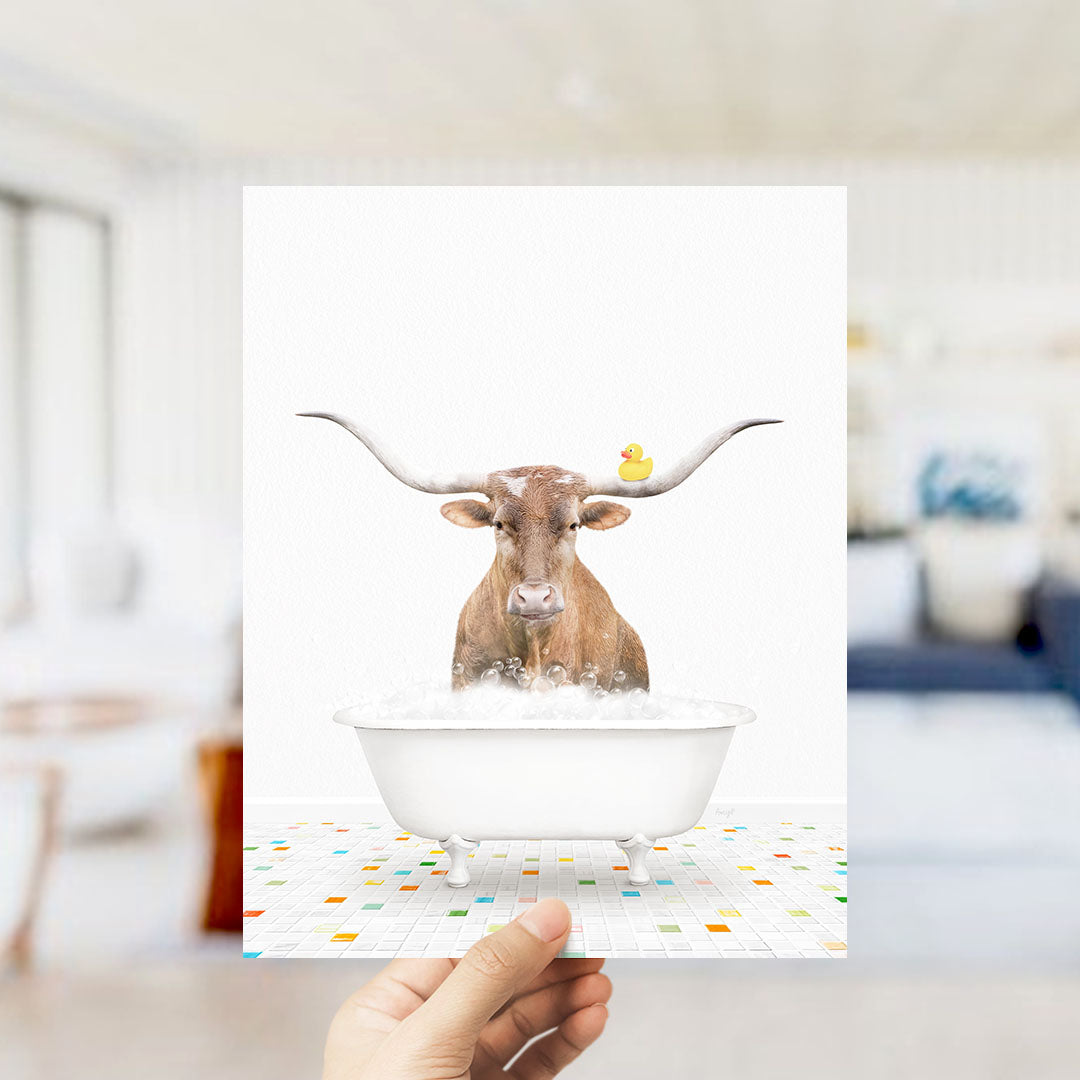 A hand is holding up a framed image of a brown cow with large horns sitting in a white bathtub filled with bubbles, with a rubber duck floating on the water.