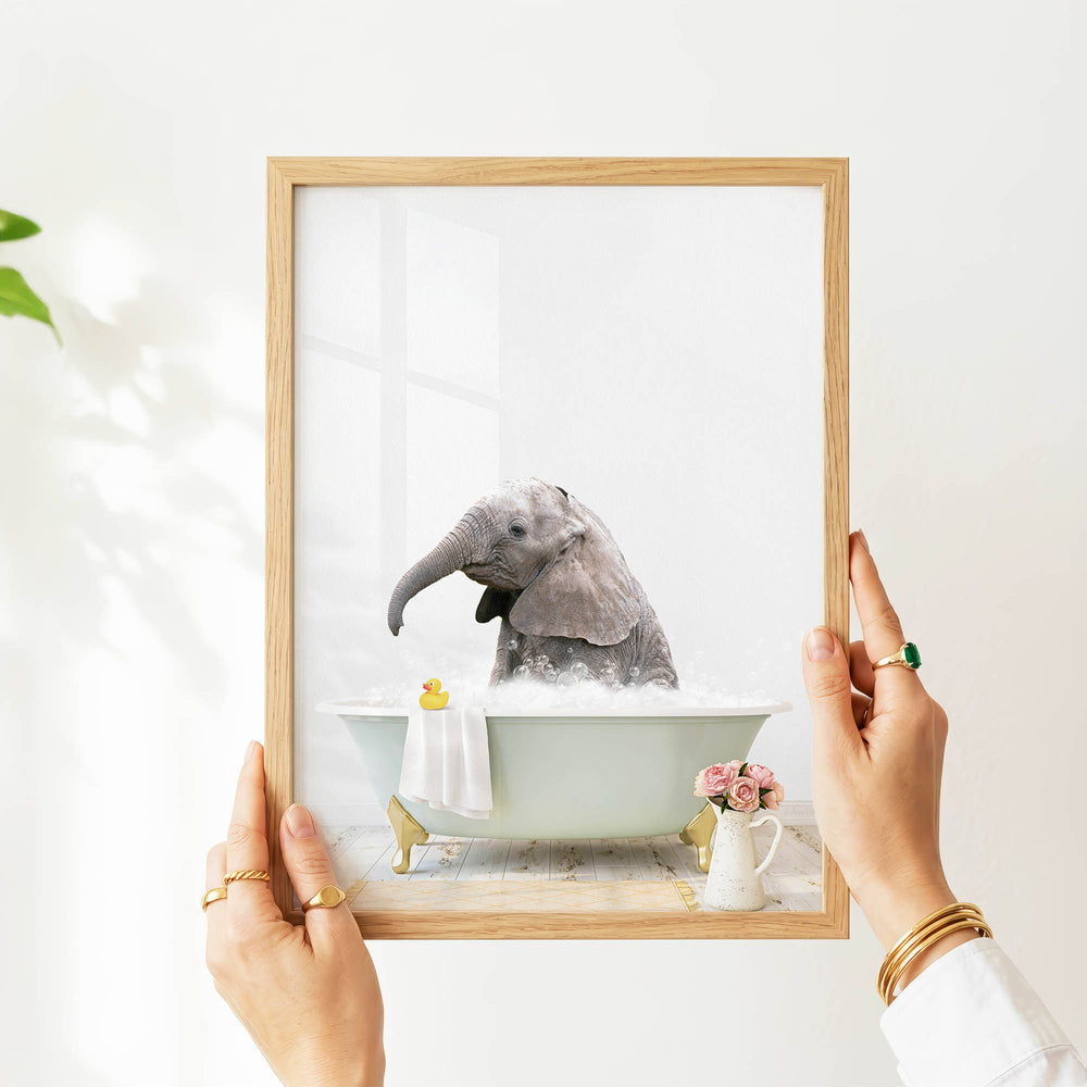 A person's hands are holding a framed picture of a baby elephant sitting in a bathtub filled with water and bubbles, with a vase of flowers and a yellow rubber duck nearby.