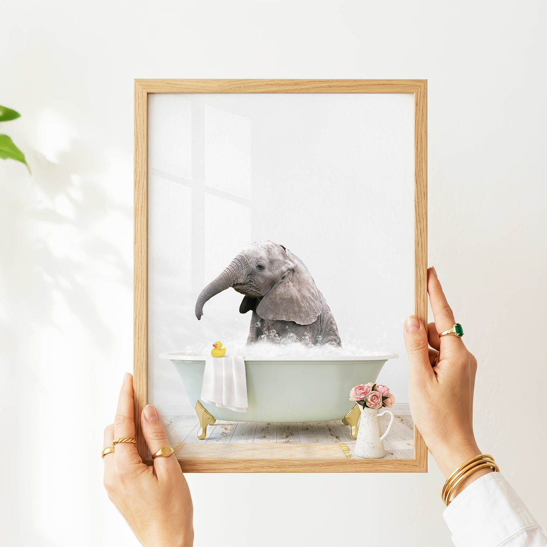 A person's hands are holding a framed picture of a baby elephant sitting in a bathtub filled with water and bubbles, with a vase of flowers and a yellow rubber duck nearby.