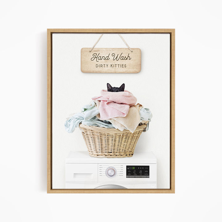 A wooden sign with the text "Hand Wash Dirty Kitties" hangs above a wicker basket filled with clothes, with a black cat peeking out from the basket.