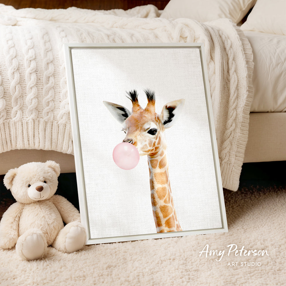 A framed image of a giraffe blowing a bubble with a pink bubble gum bubble is displayed on a white bedspread, accompanied by a teddy bear.