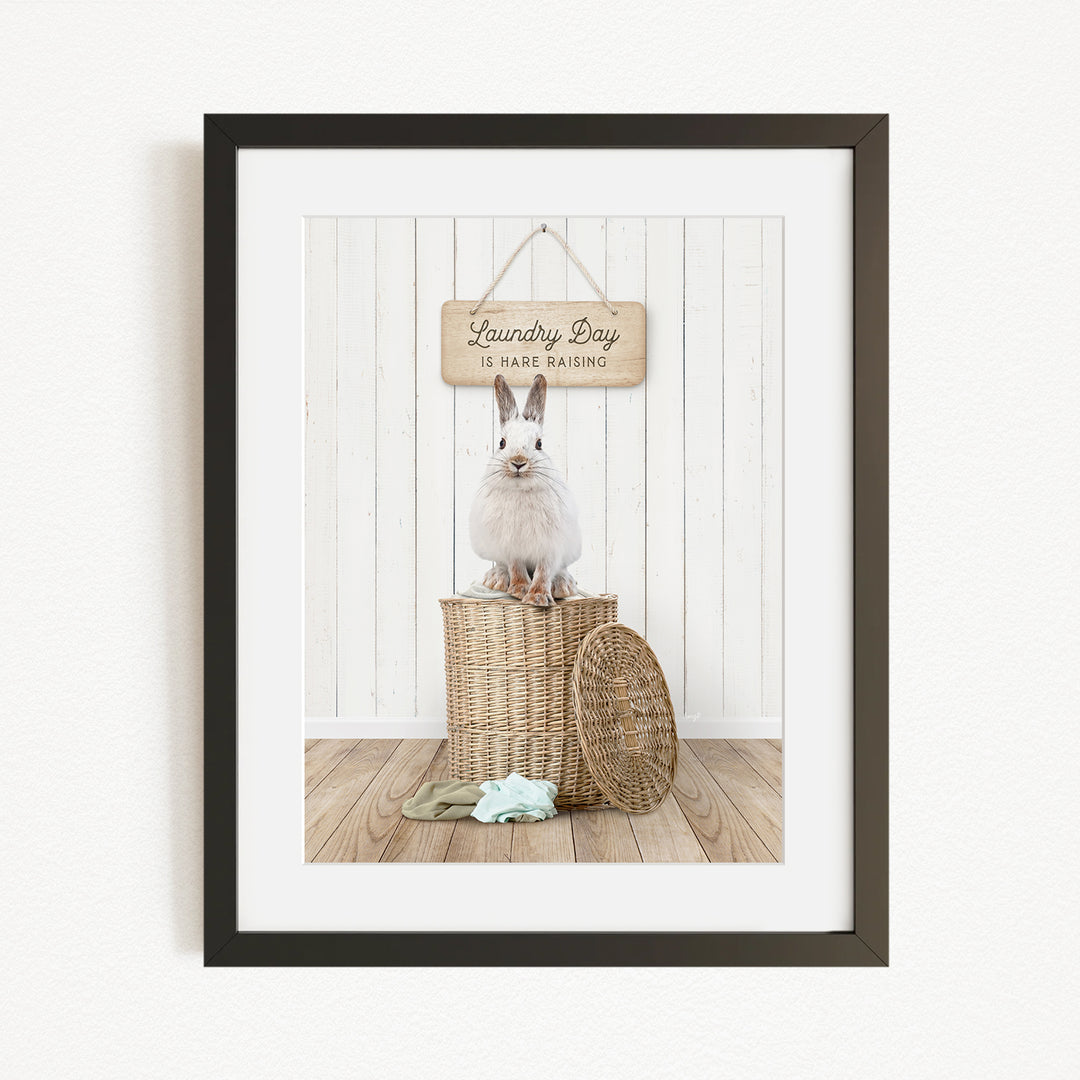 A white rabbit sits atop a wicker basket in a room with a wooden wall and a hanging sign that reads "Laundry Day is hard washing".