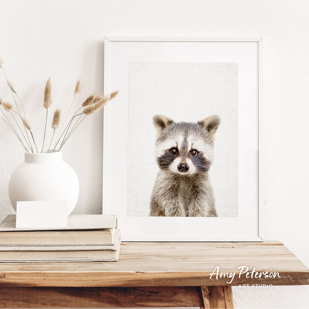 a raccoon in a white frame, sitting on a wooden shelf next to a white vase with dried flowers.