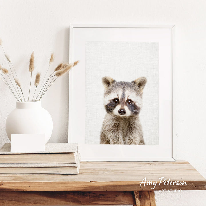a raccoon in a white frame, sitting on a wooden shelf next to a white vase with dried flowers.