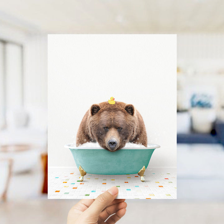 A hand holds up a framed image of a brown bear sitting in a bathtub filled with bubbles, with a yellow rubber duck floating on its head.