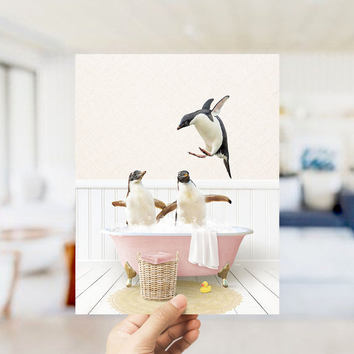 A hand holds up a framed image depicting two penguins sitting in a pink bathtub, with one penguin flying towards the other.