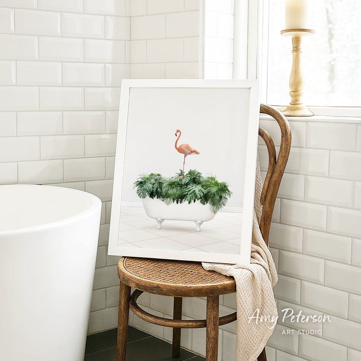 A framed image of a pink flamingo standing on a plant in a white bathtub, placed on a wooden chair in a bathroom setting.