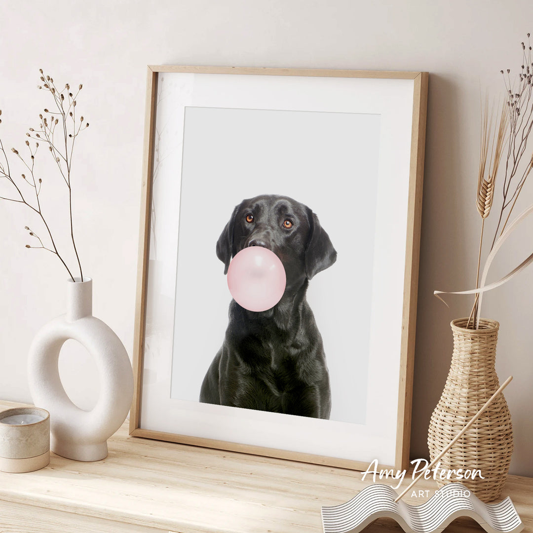 A framed photograph of a black dog blowing a bubble with a pink bubble gum bubble, accompanied by two vases and a woven basket on a wooden surface.