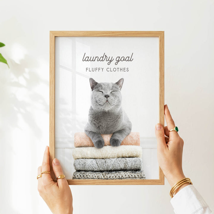 A person is holding up a framed image of a gray cat sitting on a stack of clothes, with the text "laundry goal" and "fluffy clothes" written above the cat.