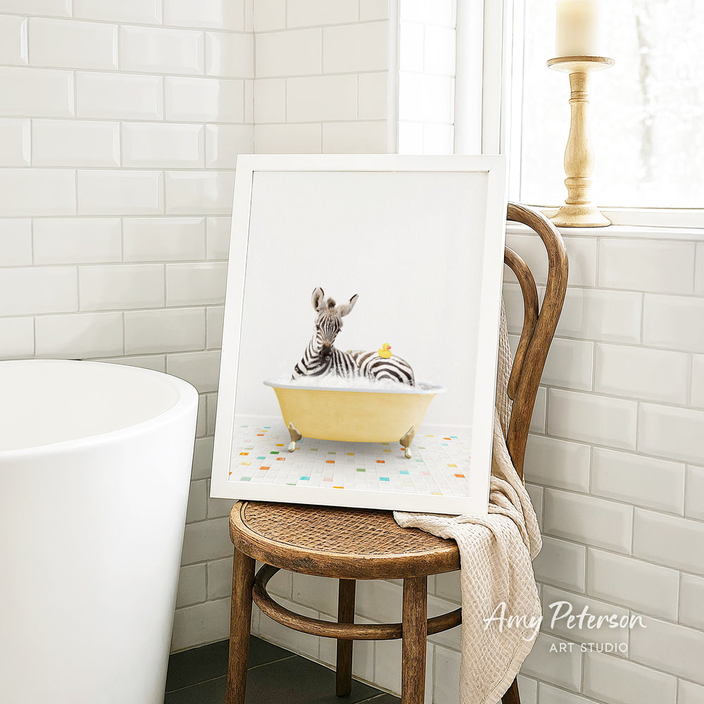 A framed image of a zebra sitting in a yellow bathtub, with a towel draped over the back of a wooden chair in a bathroom setting.