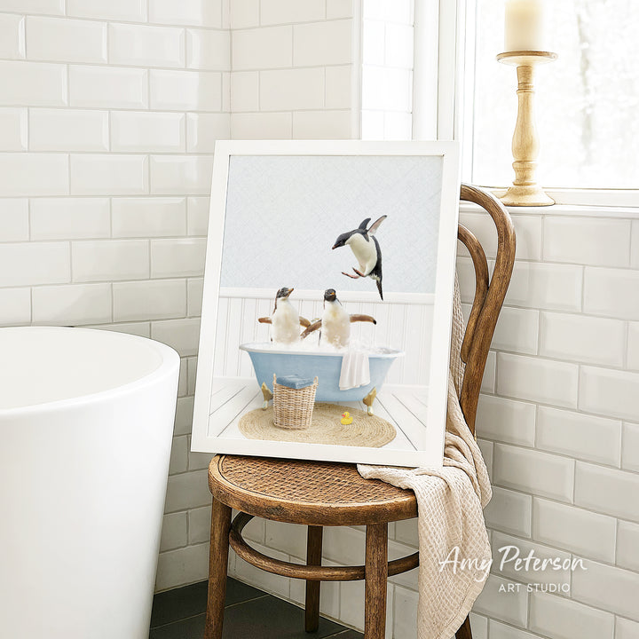 A framed image of two penguins sitting in a bathtub, surrounded by a basket of bath toys, is displayed on a wooden chair in a bathroom setting.