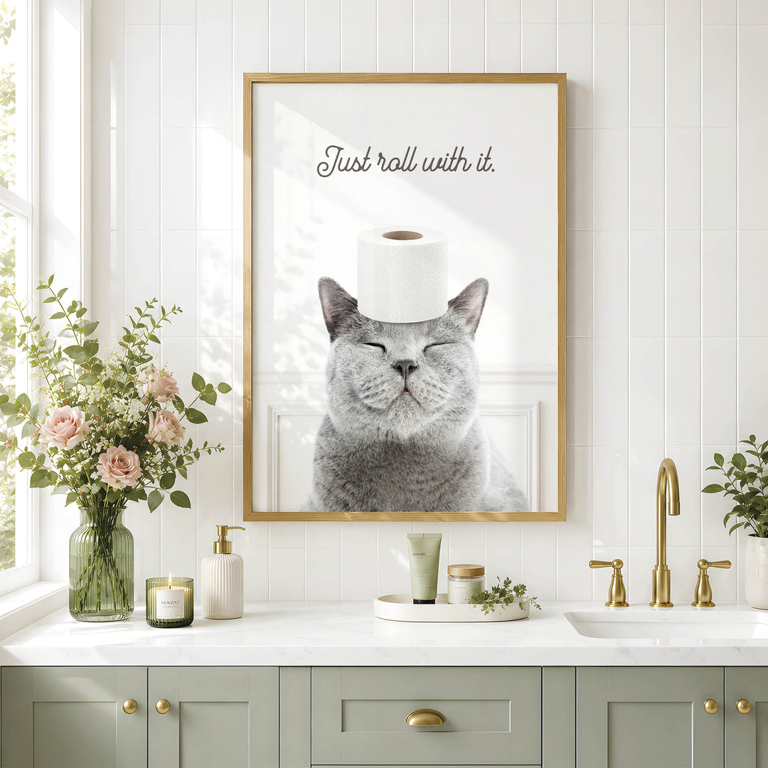 a gray cat with its eyes closed, wearing a roll of toilet paper on its head, and sitting in front of a framed picture on a bathroom wall.